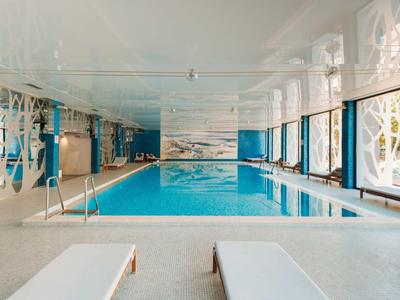 Piscina cubierta luminosa con grandes ventanales, tumbonas y diseño moderno en un hotel.