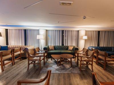 Amplia zona de estar en un hotel con sillas de madera, sofás y varias lámparas sobre suelo de madera clara.