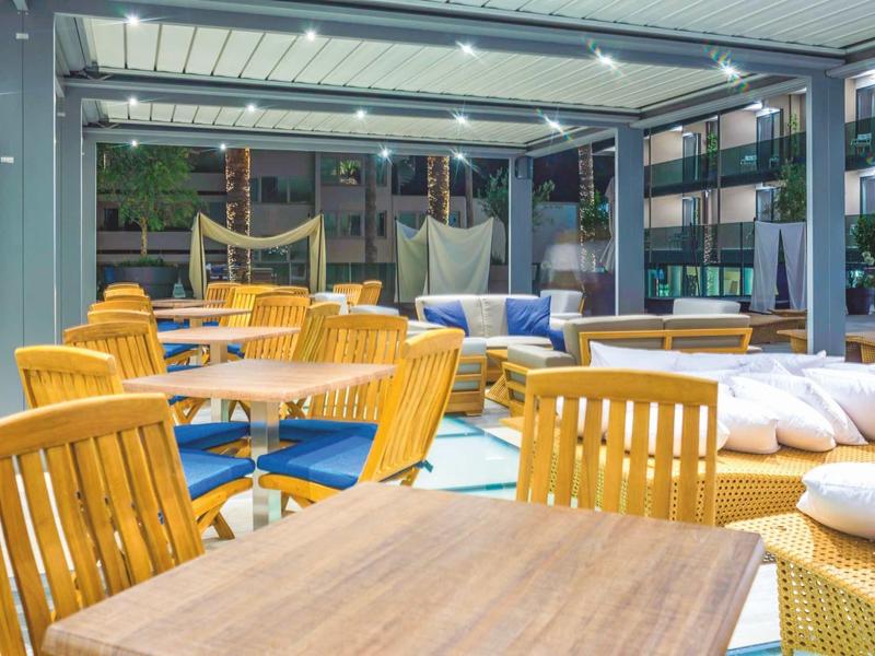 Área exterior moderna de una terraza de hotel con mesas de madera, sillas y cojines lounge bajo un techo de vidrio.