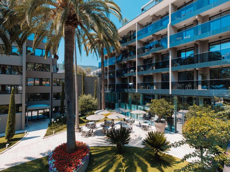 Complejo hotelero moderno de varios pisos, balcones de vidrio y un patio interior con palmeras y vegetación.