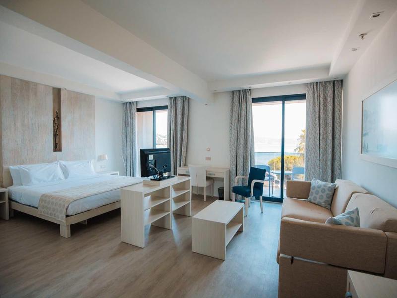 Habitación de hotel luminosa con cama doble, sofá, mesa y balcón con vista al mar.