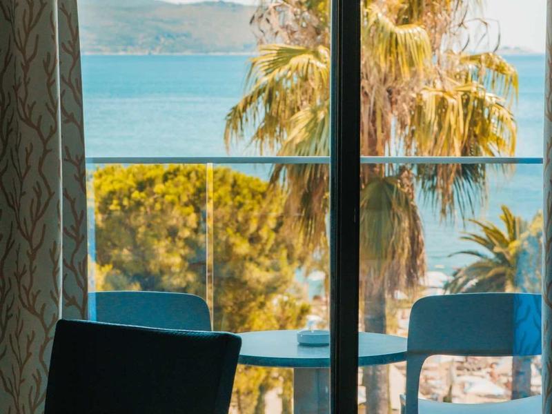 Vista desde una habitación con balcón de palmeras y el mar en un día soleado.
