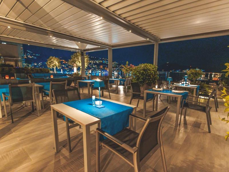 Terraza de restaurante nocturna con mesas y sillas, iluminada y con vista a luces de la ciudad.