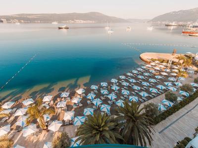 Strand mit vielen Sonnenschirmen am blauen Meer, Berge im Hintergrund und klarer Himmel.