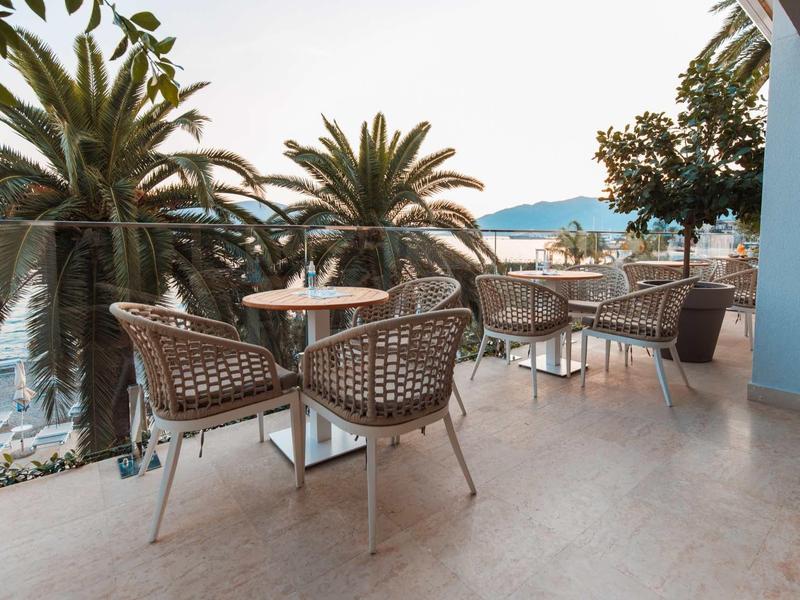 Terras met ronde tafels en stoelen, palmbomen en uitzicht op zee bij zonsondergang.