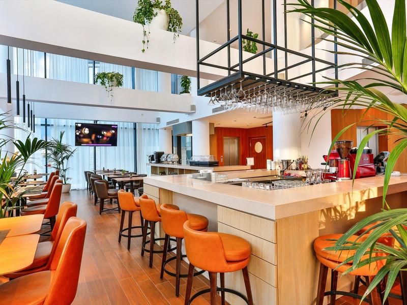 Modern hotel bar area with orange chairs and green plants in a bright space.