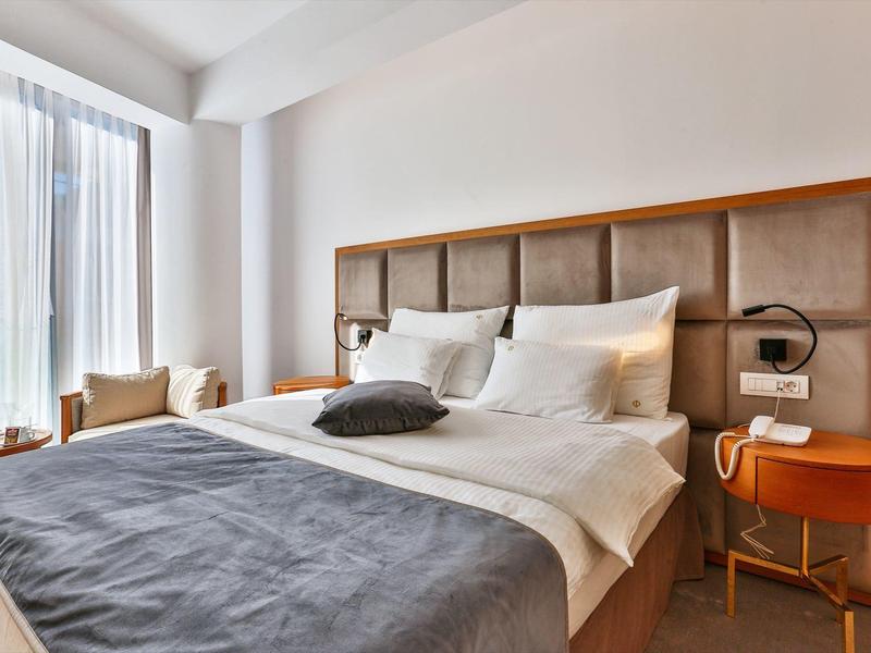 Modern hotel room with large bed, sofa, and daylight through large windows.