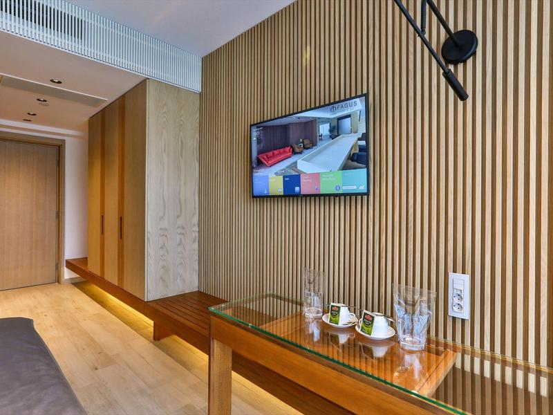Modern hotel room with wooden slat wall, wall-mounted TV, and glass table with water bottle and glasses.