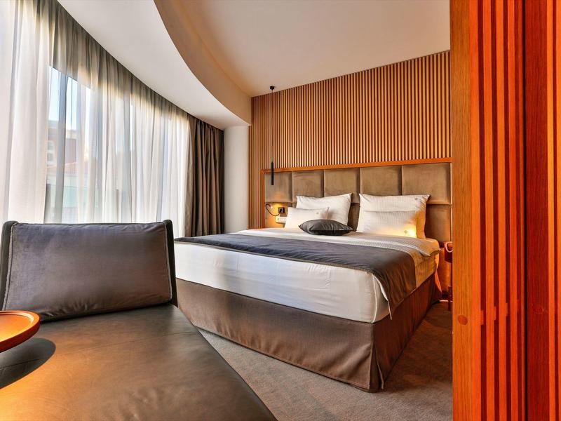 Modern hotel room with large bed, wooden wall, armchair, and curtains on the window.