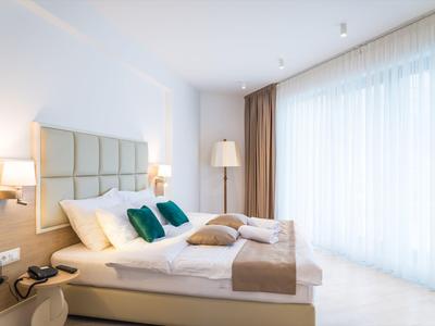 Modernes Hotelzimmer mit großem Bett, Kissen und Vorhängen vor bodentiefem Fenster.