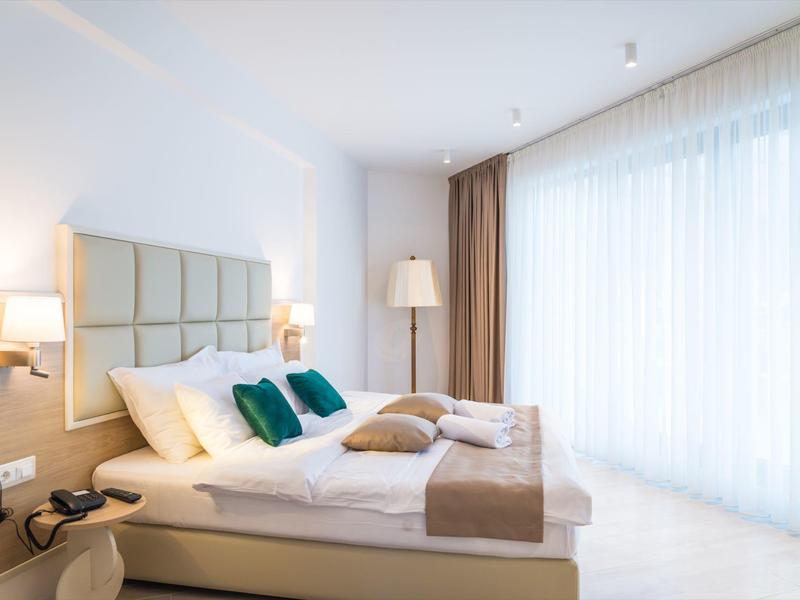 Bright hotel room with double bed, green pillows, and white curtains.