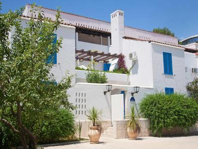 Casa vacacional blanca con ventanas azules, rodeada de arbustos y árboles bajo un cielo azul.