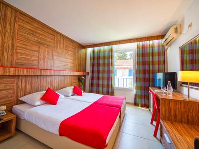 Habitación de hotel luminosa con dos camas, acentos rojos y una gran ventana con cortinas coloridas.