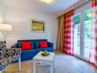 Habitación de hotel luminosa con sofá azul, cojines coloridos, sillón y ventana grande con cortinas.