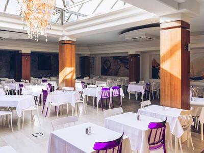 Großer, heller Speiseraum mit Tischen, weißen Tischdecken und lila Stühlen unter einem Glasdach.