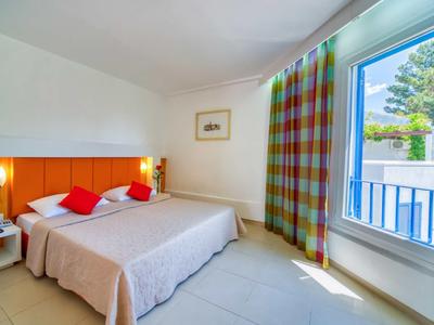 Habitación de hotel luminosa con cama doble grande, cojines rojos y vista al balcón con jardín.