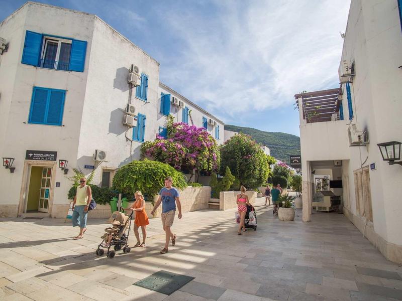 Calle peatonal con edificios blancos y ventanas azules, personas caminando y árboles floridos.