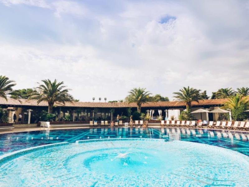 Gran piscina al aire libre con mosaicos azules, rodeada de tumbonas y palmeras bajo el cielo azul.