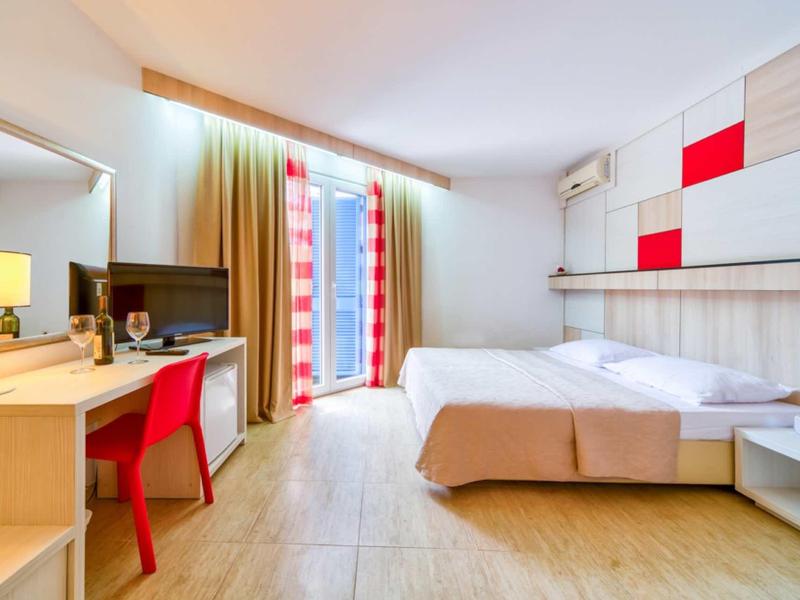 Habitación de hotel luminosa con cama doble, escritorio, silla roja y puerta balcón con cortinas.