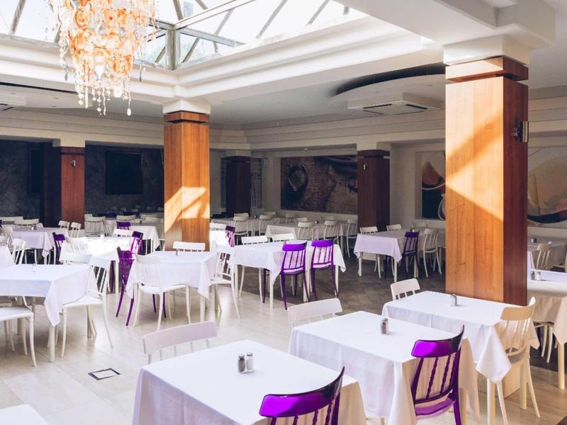 Restaurante de hotel luminoso con mesas blancas, sillas moradas y un gran techo de vidrio.