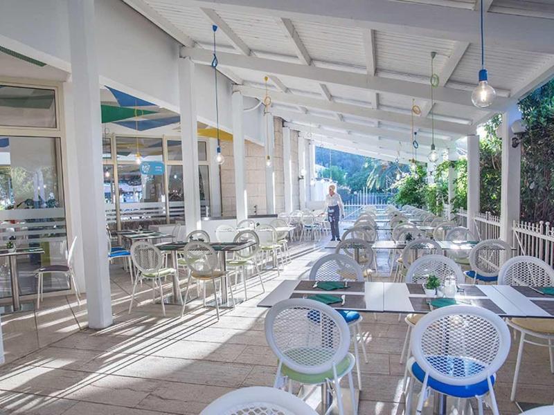 Área exterior moderna de un hotel con mesas y sillas blancas bajo una pérgola a la luz del día.