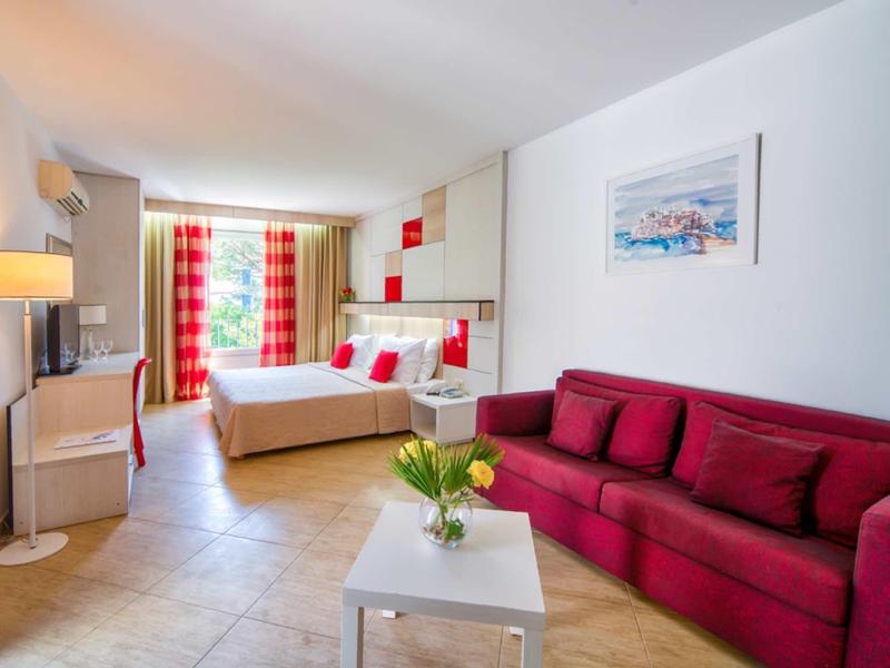 Habitación de hotel luminosa con sofá rojo, cama doble y balcón con cortinas en tonos rojos y verdes.