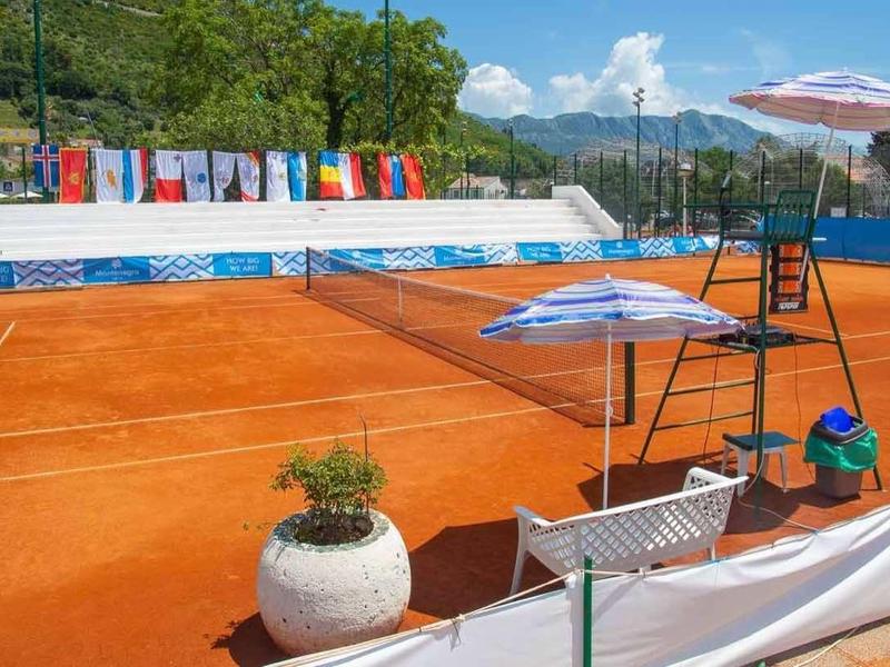 Cancha de tenis al aire libre con polvo naranja, asientos y sombrillas al lado de la red.