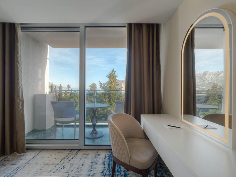 Camera d'hotel luminosa con scrivania, sedia e balcone con vista su montagne e lago.