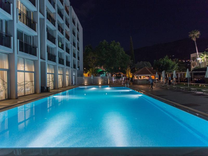 Piscina dell'hotel illuminata di notte con edifici moderni e palme sullo sfondo.