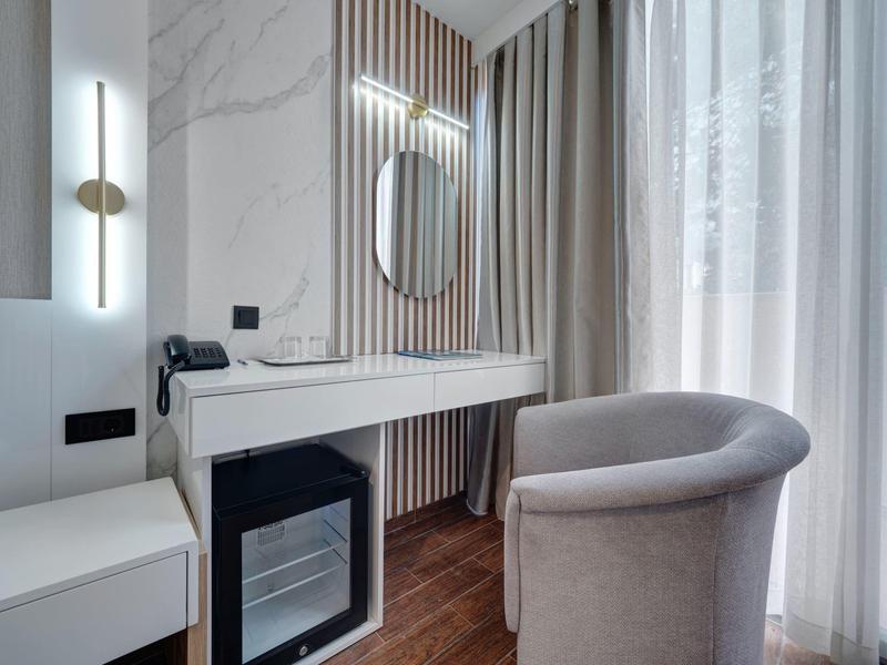 Scrivania moderna in camera d'hotel con sedia, minibar e specchio illuminato accanto a finestra con tende.