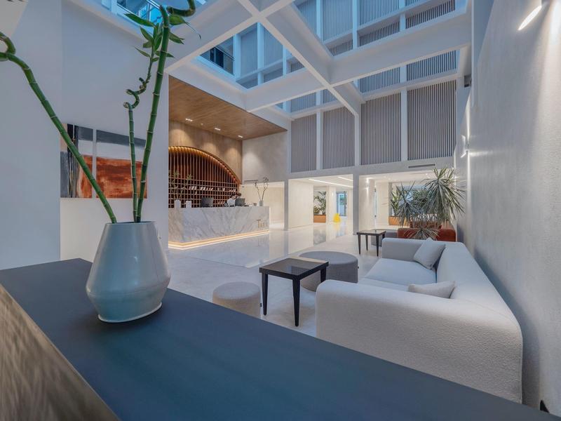 Lounge dell'hotel luminoso con divano bianco, tavoli e ampio atrio con bar sullo sfondo.