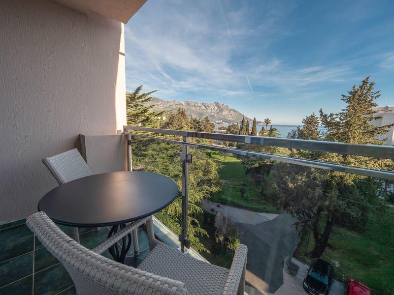 Balcone con tavolo e sedie che si affaccia su giardino e montagne sotto un cielo azzurro.