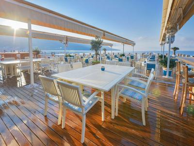 Große Terrasse mit weißen Tischen und Stühlen, Holzbohlenboden, Blick aufs Meer, sonniger Himmel.