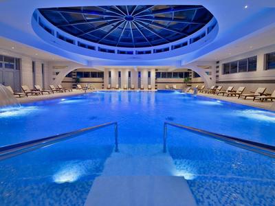 Großer, beleuchteter Hallenbadbereich mit blauer Wasserfläche, Liegestühlen und rundem Glasdach.