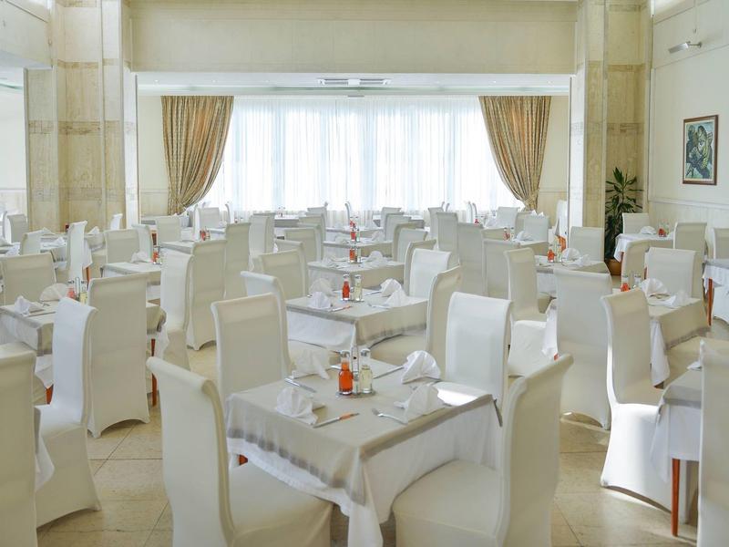 Helles Restaurant mit weißen Stühlen, Tischen, großen Fenstern und hellen Vorhängen.