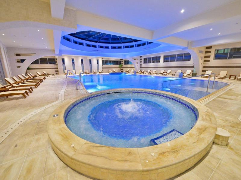 Großer Hallenbadbereich mit rundem Whirlpool, mehreren Liegestühlen und blauen Wasserelementen.