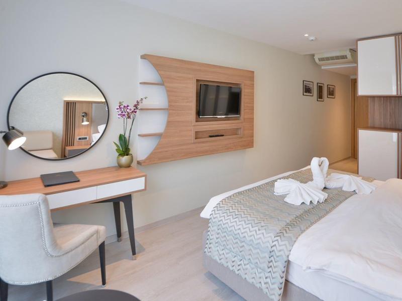 Modernes Hotelzimmer mit hellem Holzboden, Schreibtisch, runden Spiegel und Fernseher an Holzpaneel.