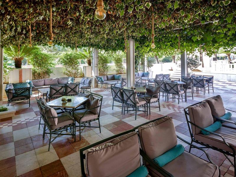 Überdachte Terrasse mit grünen Pflanzen, Sitzgruppen aus Metall und gepolsterten Stühlen.