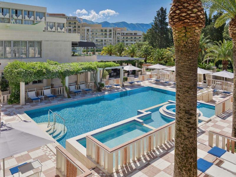 Moderner Außenpool mit blauer Wasserfläche, Liegestühlen, Palmen und Hotelgebäuden im Hintergrund.