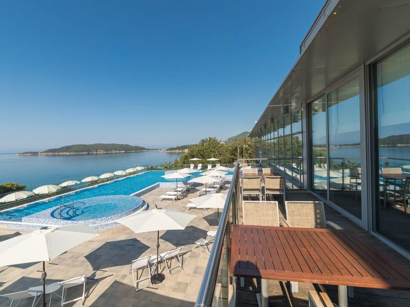 Große Terrasse mit Tischen und Stühlen neben Pool, Sonnenschirmen und Blick auf Meer und Insel.