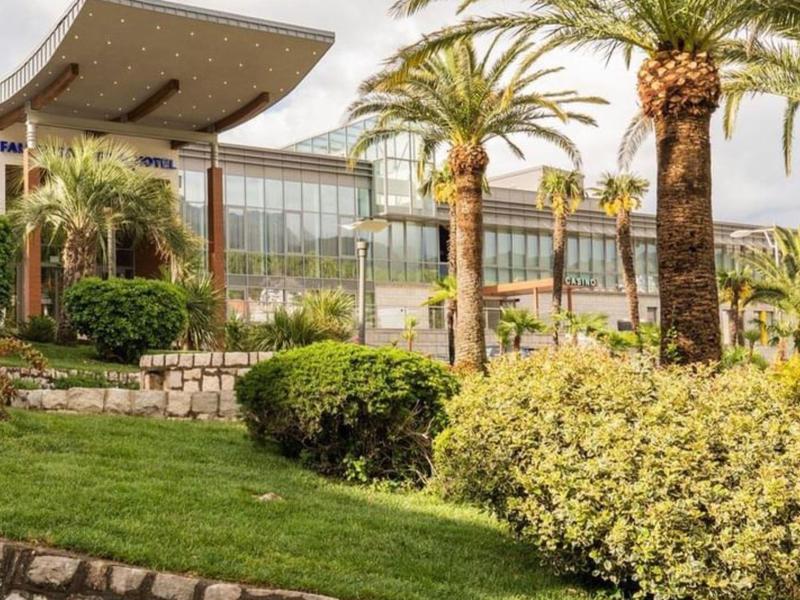 Grüner Garten mit Palmen und Buschwerk vor modernem Glasgebäude unter bewölktem Himmel.