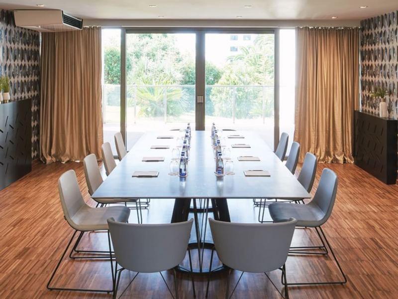 Moderner Konferenzraum mit langem Tisch, grauen Stühlen, Holzboden und großen Fensterfronten.