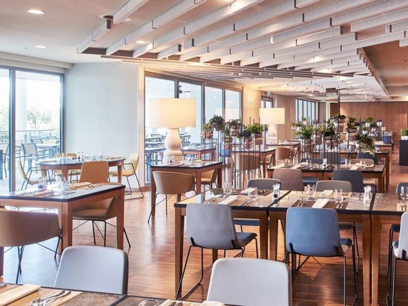 Moderner, heller Restaurantraum mit Holztischen, Stühlen und großen Fenstern für Tageslicht.