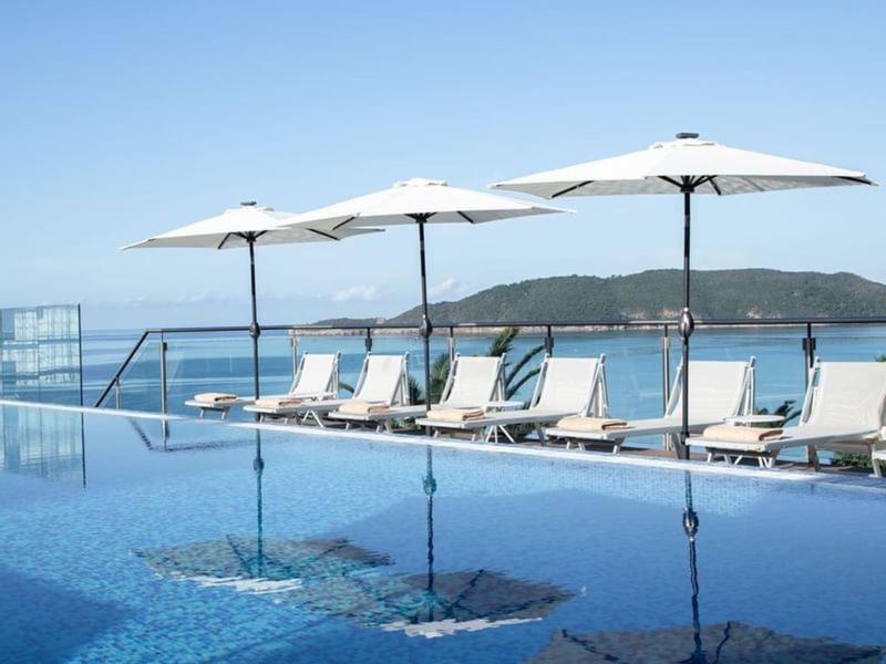 Infinity-Pool mit Liegestühlen und weißen Sonnenschirmen vor blauem Meer und grünem Hügel.