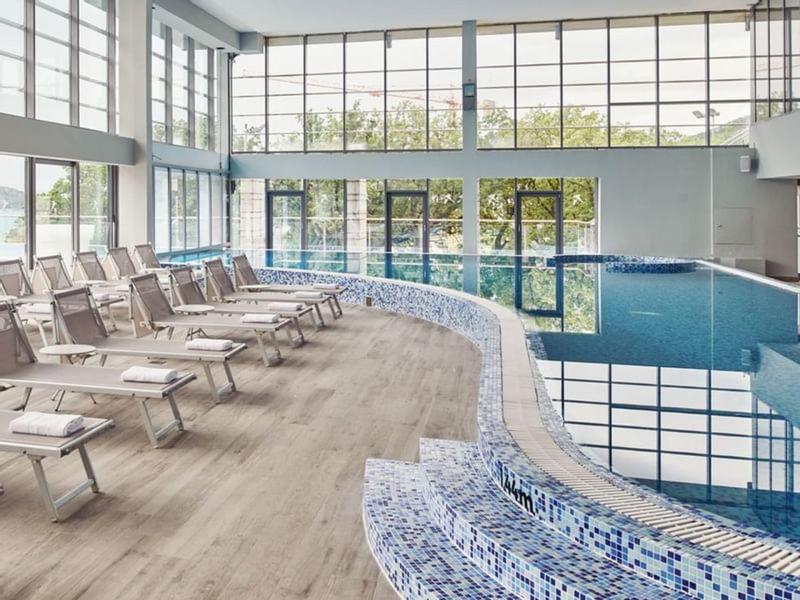 Innenbereich mit großem Schwimmbecken, blauen Fliesen und liegenden Stühlen an hellem Holzboden.