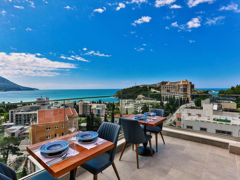 Pohled z terasy s stolky a židlemi na moře a okolní budovy pod modrou oblohou.