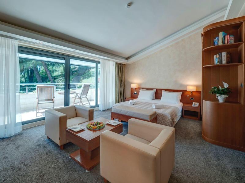 Habitación de hotel luminosa con cama doble, sillones beige y vistas a la terraza con vegetación.