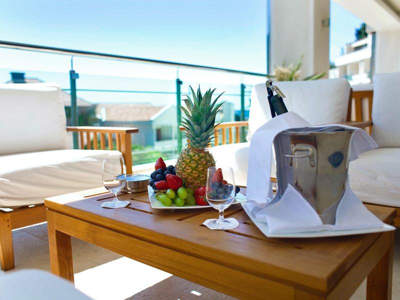 Balcón moderno con mesa de madera, vino, cubitera y frutas tropicales con vista al mar al fondo.