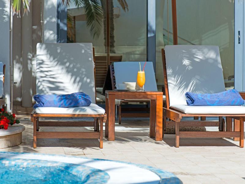 Dos tumbonas con toallas azules junto a una mesa con bebidas junto a la piscina frente a una pared de cristal.