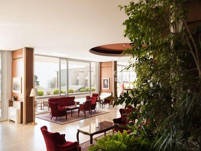 Area moderna della hall dell'hotel con grandi finestre, poltrone rosse e piante verdi all'interno.